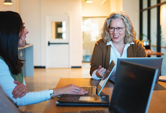 Two employees sitting at a table in an office workspace, collaborating with laptops open and engaged in conversation.