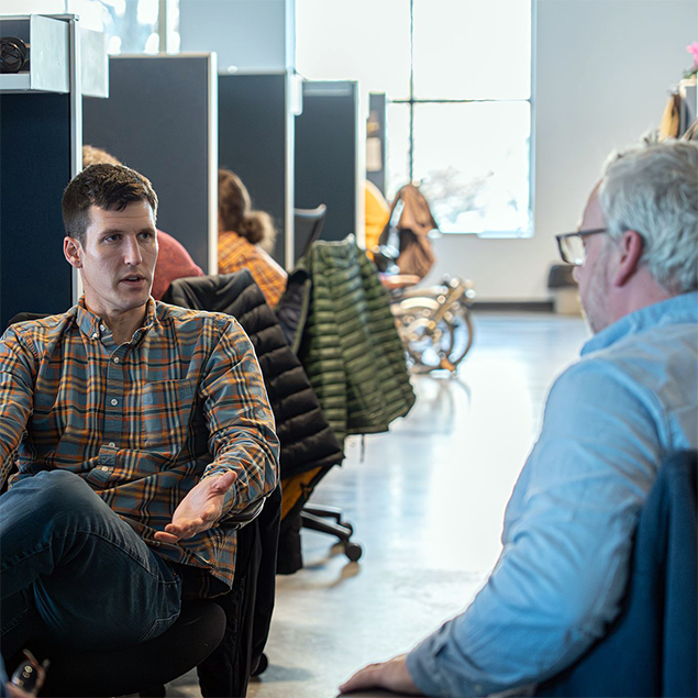 Two employees seated in an open office workspace engaged in conversation, with coworkers and desks visible in the background.