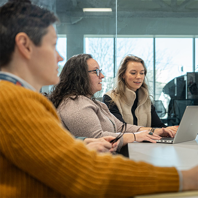 Photograph of three Onpoint team members seated at a conference table in a modern office with large windows, engaged in a meeting or collaborative work. One person in a tan fleece vest is using a laptop.