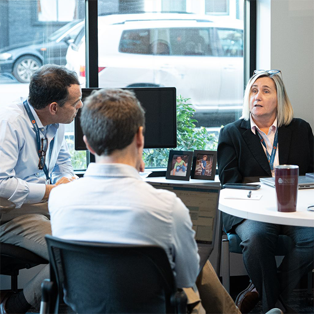 Photograph of three professionals engaged in a meeting around a round table in an office with a large window showing parked cars outside. The setting includes laptops, a tumbler, framed photos on a windowsill, and a monitor, indicating a collaborative work discussion.