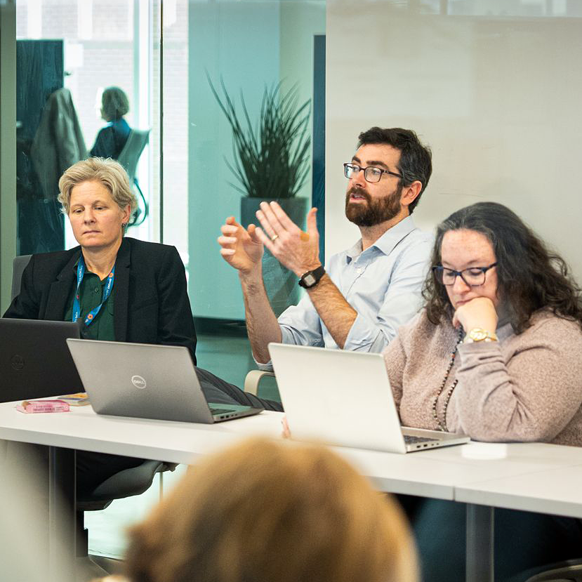 Photograph of a business meeting showing three Onpoint team members seated at a conference table with laptops, engaged in discussion. The setting includes a modern office with glass walls, a visible plant, and a person working at their desk in the background, highlighting a collaborative work environment.