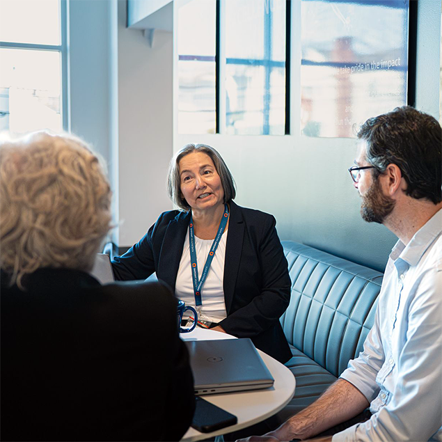 Photograph of three professionals engaged in a discussion around a small round table in a modern office setting. Key elements include a laptop, coffee mugs, and large windows providing natural light, highlighting a collaborative work environment.