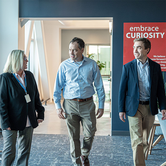 Photograph of three business professionals walking and conversing in a modern office hallway with large windows and natural light. A red wall sign with white text displays the company value of "embrace curiosity," emphasizing a workplace culture focused on innovation and learning.