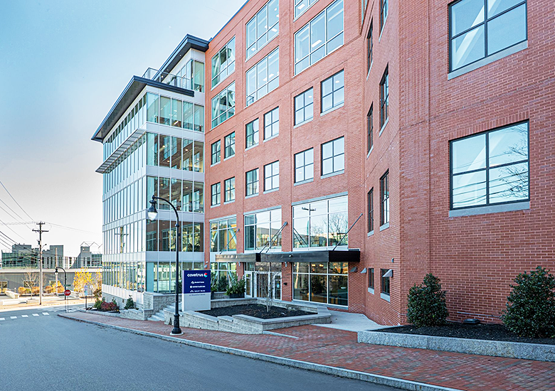 Photograph of a modern commercial building with red brick and glass facades along a quiet street. The scene includes a sidewalk with small shrubs, a street lamp, and a sign indicating company names, highlighting an urban business setting in the Old Port district in Portland, Maine.