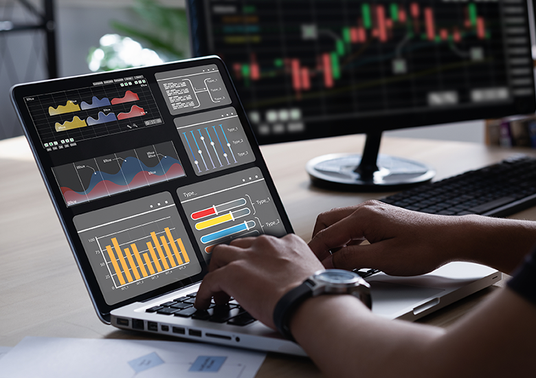 Photograph of a laptop screen displaying multiple data visualizations, including bar charts, line graphs, and slider controls, used for data analysis or monitoring. A second monitor in the background shows a candlestick stock chart, indicating trend analysis.