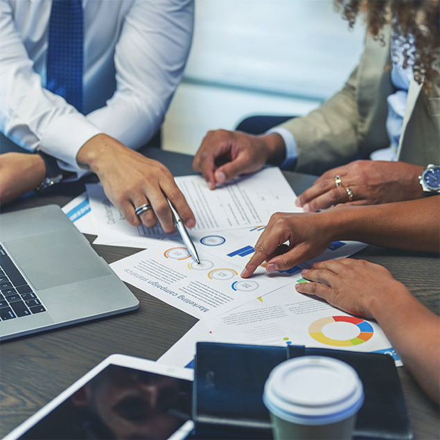 Photograph of a business meeting showing three people discussing printed charts and graphs on a table, including pie charts and circular diagrams with color-coded sections. Key elements include a laptop, tablet, and coffee cup, indicating a collaborative work session focused on data analysis and strategy planning.