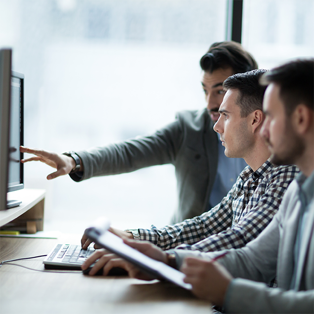 Photograph of three people collaborating at a desk with a computer, focusing on one person pointing at the monitor while another types on the keyboard and a third takes notes on a clipboard. The setting suggests a collaborative work environment emphasizing teamwork and information sharing.