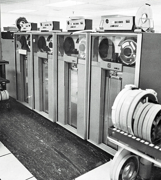 Black and white photograph of a row of vintage mainframe computer tape drives with large reels of magnetic tape, showcasing early data storage technology.