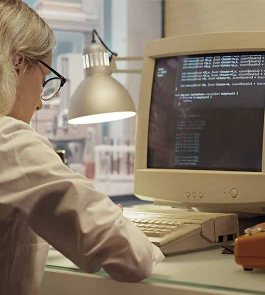 Photograph of a person working at a desk with an old-style CRT monitor displaying lines of computer code in an office setting.