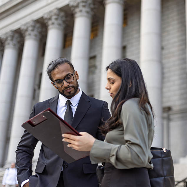 Photograph of two professionals standing outside a government building with large columns, engaged in discussion while reviewing unseen materials on a clipboard. The man wears a suit and tie, and the woman wears a blouse, indicating a professional context.