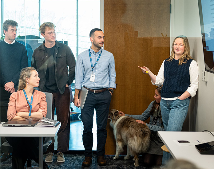 Photograph of Onpoint staff listening to a presentation from a colleague in a glass-walled conference room. The image features six people and a dog. One person stands and gestures while others listen, with a laptop and notebook visible on tables, and a wooden door and glass window in background.