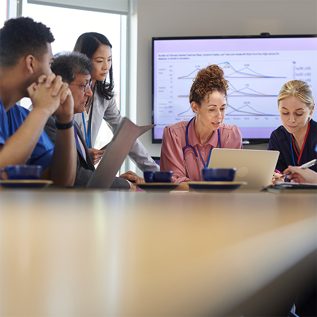 Photograph of a group of medical professionals in a meeting room discussing data displayed on a large screen showing multiple line graphs with labeled axes and color-coded lines. The graphs likely represent trends in patient metrics or hospital performance, with key markers indicating significant changes or milestones over time.