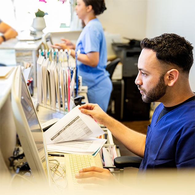 Photograph showing healthcare staff working at a medical office reception desk with computers, paperwork, and hanging file folders. Focus is on a male worker reviewing intake and claims documents while a female worker interacts with a computer and colleague in the background, highlighting administrative tasks in a clinical setting.