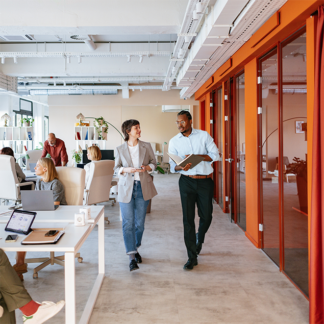 Photograph of a modern open-plan office with employees engaged in various work activities, including two individuals walking and conversing in the foreground. The space features white ceilings with exposed ducts, orange-framed glass office rooms on the right, and desks with laptops and office supplies on the left, highlighting a collaborative and dynamic work environment.