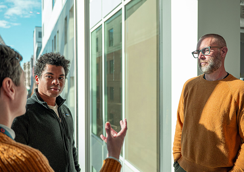 Photograph of three Onpoint staff members engaged in a conversation near large windows in a modern building with natural light illuminating the scene.