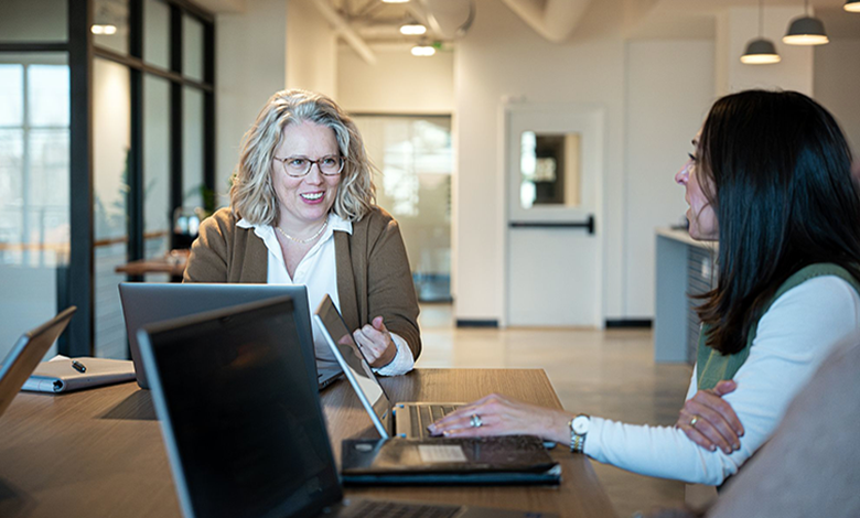 Photograph of two women engaged in a discussion at a modern office table with laptops and notebooks. The setting features large windows, contemporary lighting, and an open workspace, highlighting a collaborative work environment.