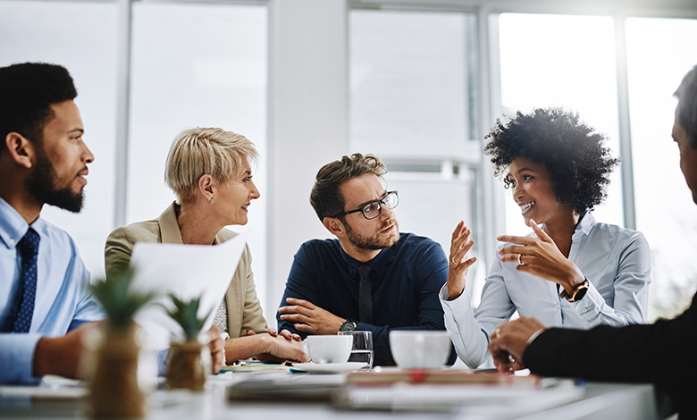 Photograph of a business meeting with five professionals engaged in discussion around a table with coffee cups and documents. The bright office setting with large windows highlights active communication and collaboration among diverse team members.