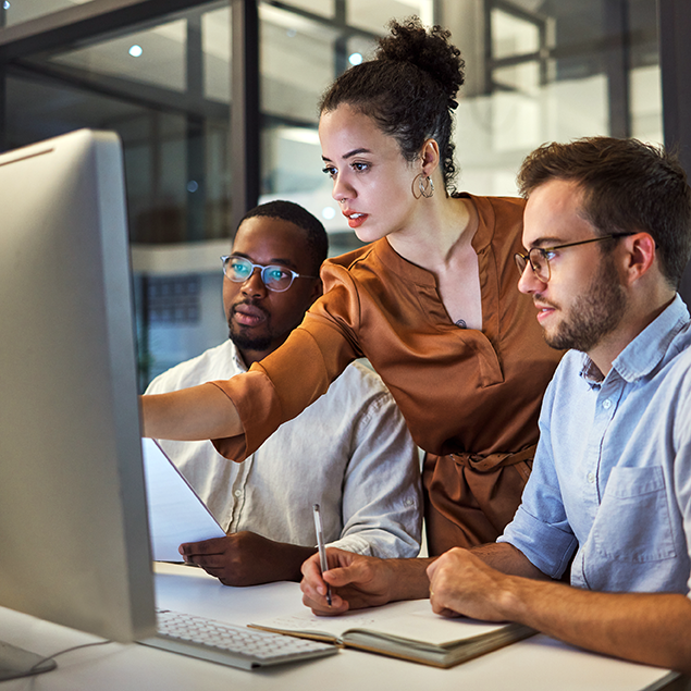 Photograph of three coworkers collaborating at a desk with a large computer monitor in a modern office setting. One person in a copper-colored blouse is pointing at the screen while the others, one holding a document and the other taking notes, attentively engage in discussion.