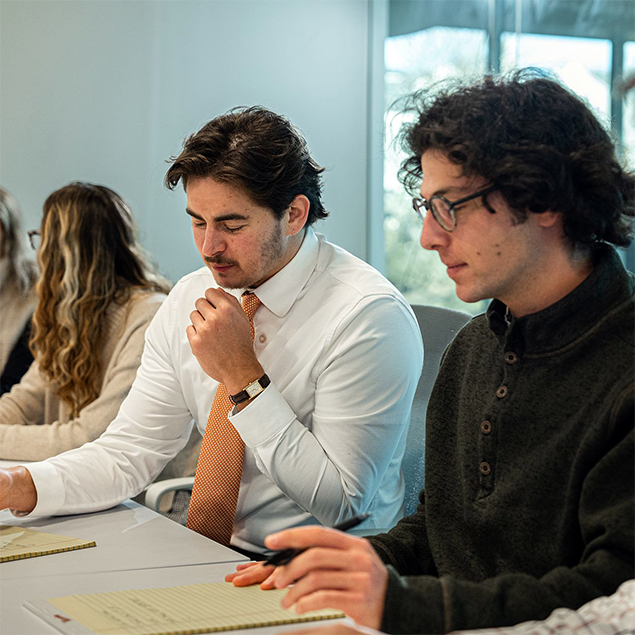 Photograph of a group of people seated at a table engaged in a meeting and discussion. Individuals are taking notes on notepads and laptops, with natural light coming through windows in the background.