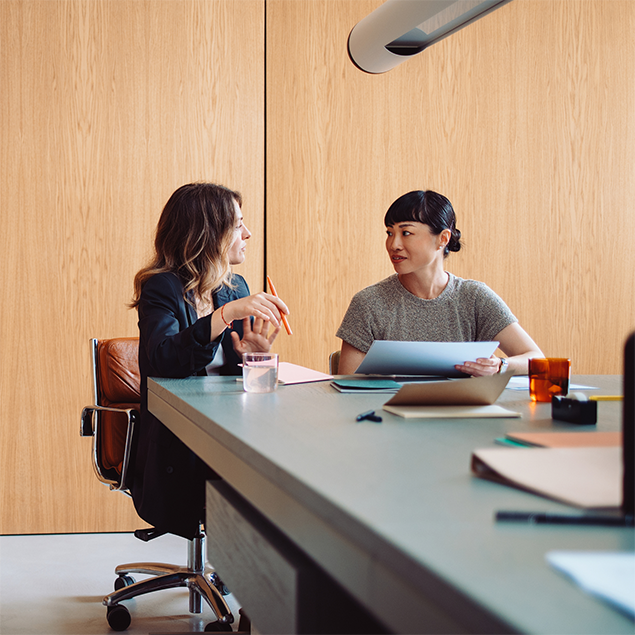 Photograph of two women sitting at a conference table in an office setting, engaged in discussion with documents and notebooks in front of them. The scene features a wooden wall background, modern office chairs, and various office supplies on the table, highlighting a collaborative work environment.