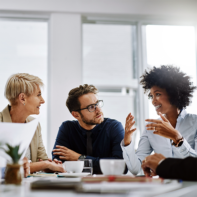 Photograph of a business meeting with three professionals engaged in discussion around a table with coffee cups and documents. The bright office setting with large windows highlights active communication and collaboration among diverse team members.