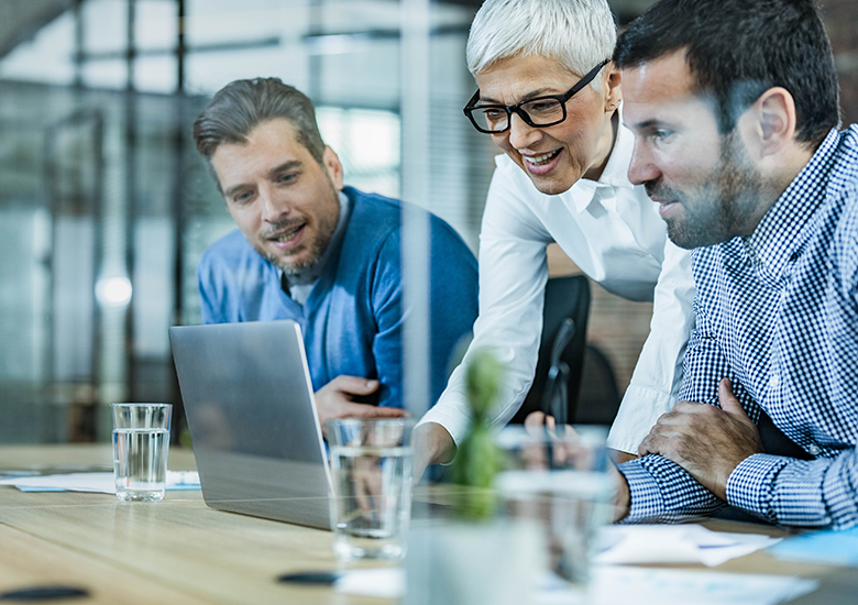 Photograph showing three business professionals collaborating around a laptop in a modern office setting. Focus on teamwork and discussion is highlighted by their attentive postures, casual business attire, and engaged expressions.