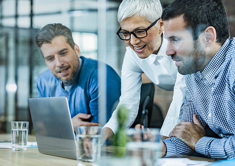 Photograph showing three business professionals collaborating around a laptop in a modern office setting. Focus on teamwork and discussion is highlighted by their attentive postures, casual business attire, and engaged expressions.
